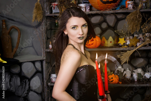 A beautiful, young witch in a black corset with burning red candles in her hands and long hair. Studio photo. Girl in suit on Halloween. A model with clean skin.