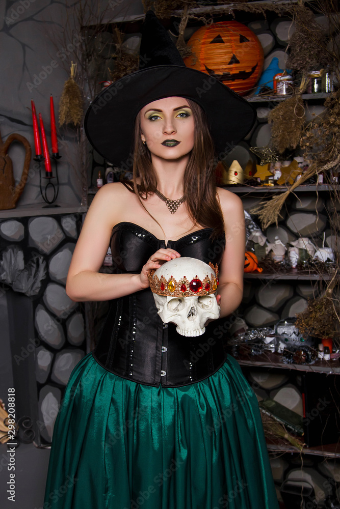 A young, beautiful girl in a green skirt, a black corset and a black hat holds a skull with a crown in her hands. Witch costume, Halloween, photo studio, model with clean skin.