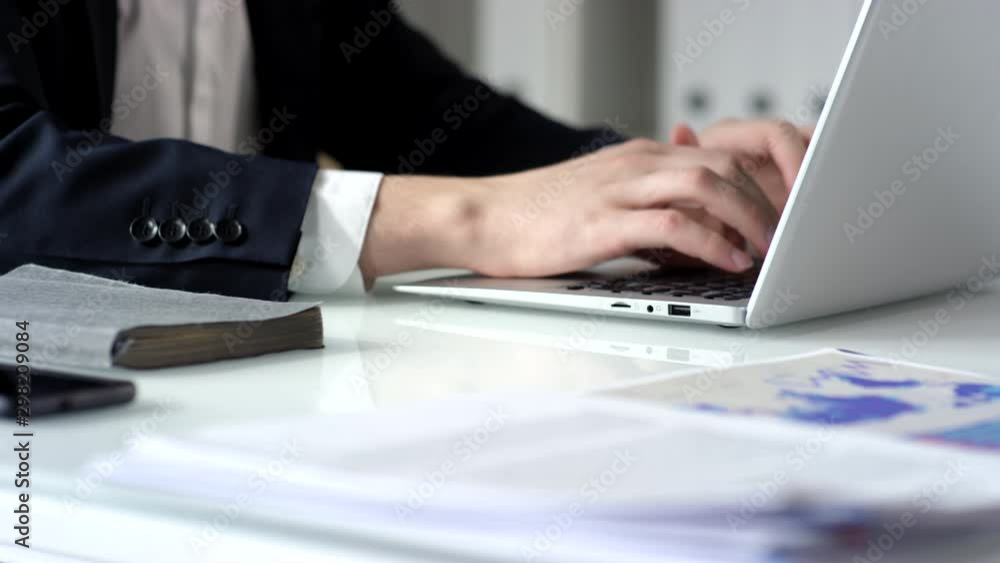 Zoom in rack focus shot of hands of unrecognizable businessman putting documents on the edge of office table and then typing on laptop Zoom in rack focus shot of hands of unrecognizable businessman putting documents on the edge of office table and then typing on laptop