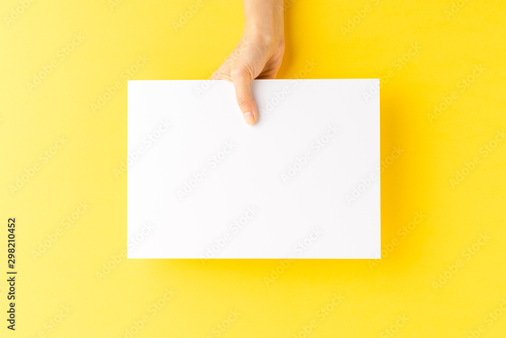 Woman’s hand showing blank a4 paper sheet on yellow background. Close up - obrazy, fototapety, plakaty 