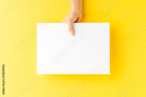 Woman’s hand showing blank a4 paper sheet on yellow background. Close up