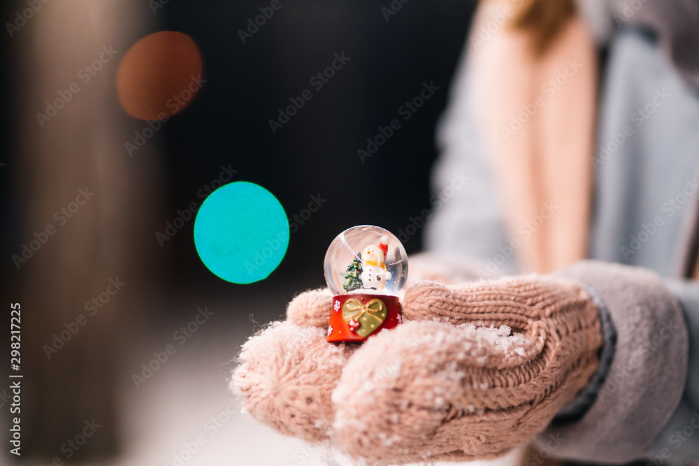New Year`s glass snow globe in hands. Happy winter time in the forest. The girl is dressed in a blue coat, in a light knitted hat, scarf and mittens. Festive garland lights. Christmas, new year.