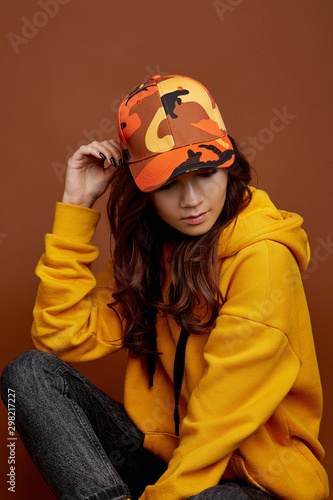 Canvas Print Cropped upward shot of a dark-haired girl wearing colorful baseball cap with camouflage print, yellow hoodie and black jeans