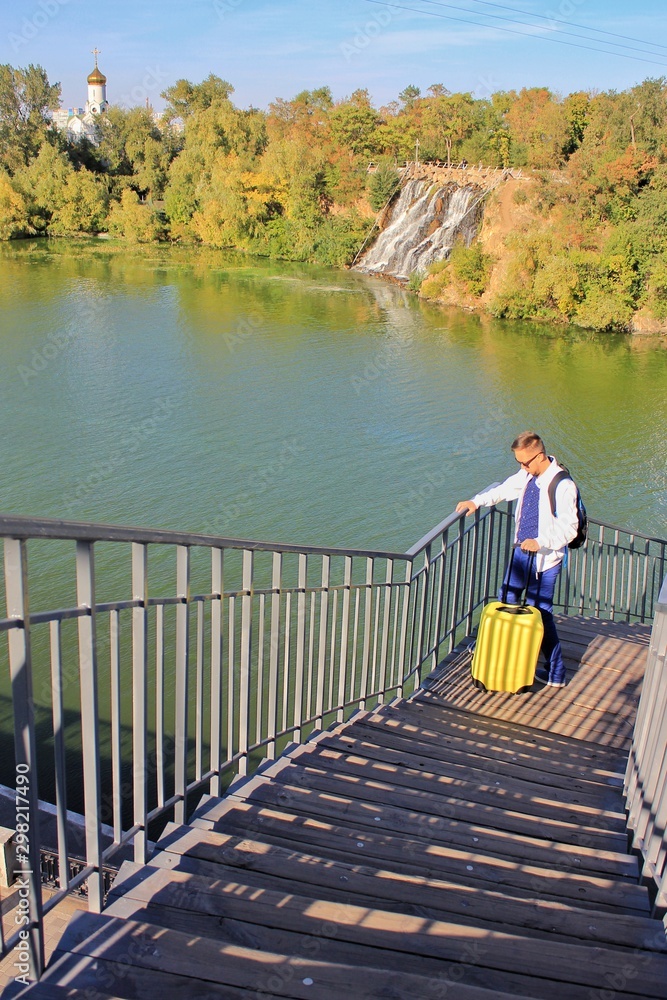 Solo traveling man with backpack and yellow suitcase with church and waterfall background 
