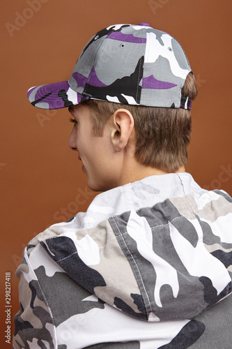 Half-turn back photo of European man with short fair hair in a camouflage hoodie, gray tee-shirt and multicolor baseball cap with a camouflage print. The man is posing on the brown background. photo