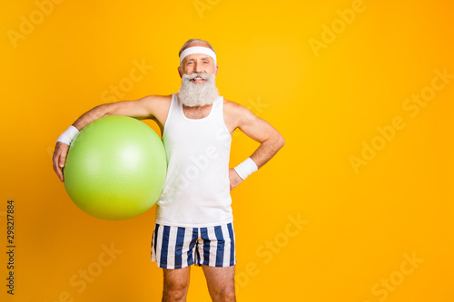 Fototapeta Naklejka Na Ścianę i Meble -  Photo of positive cheerful handsome old man holding fit ball smiling toothily with hand on waist standing confidently near empty space isolated vivid yellow color background