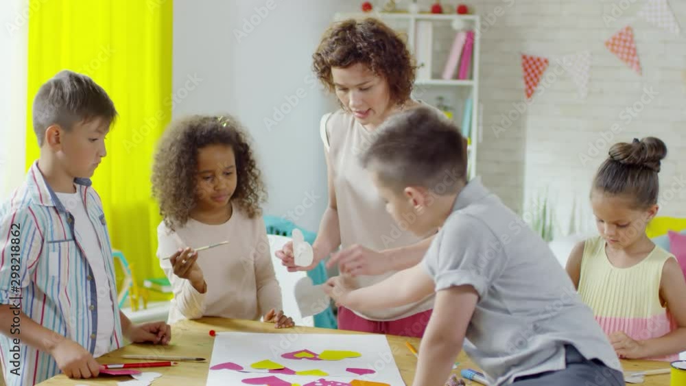 Female teacher explaining little boys and girls how to make paper collage with colorful hearts during lesson in kindergarten Female teacher explaining little boys and girls how to make paper collage with colorful hearts during lesson in kindergarten