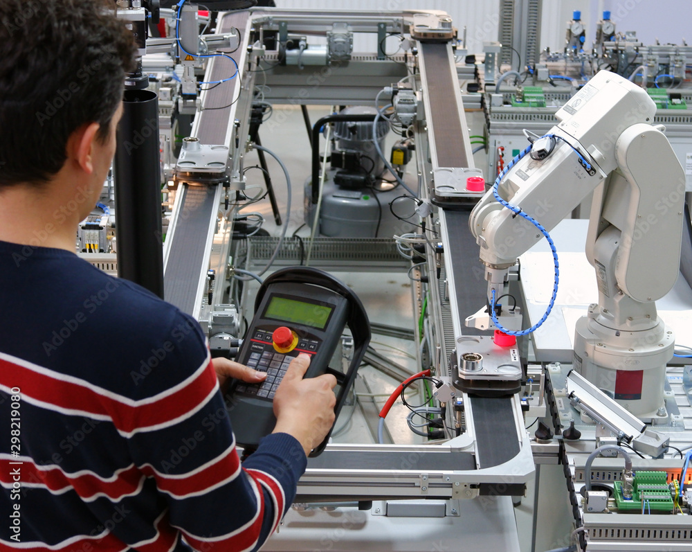 Man is programming robotic arm with teach panel which is integrated on smart factory production line. industry 4.0 automation line which is equipped with sensors and robotic arm. Selective Focus. Man is programming robotic arm with teach panel which is integrated on smart factory production line. industry 4.0 automation line which is equipped with sensors and robotic arm. Selective Focus.