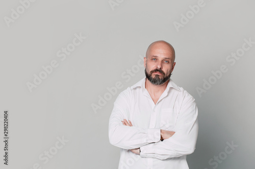 Serious middle-aged man with folded arms and a deadpan expression posing in front of a gray background with copy space © Nana_studio