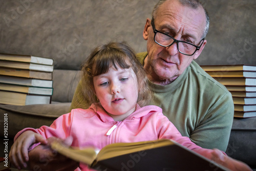 An elderly man and a little girl leaf through a thick textbook. Grandfather teaches his granddaughter to read books. Family educational concept