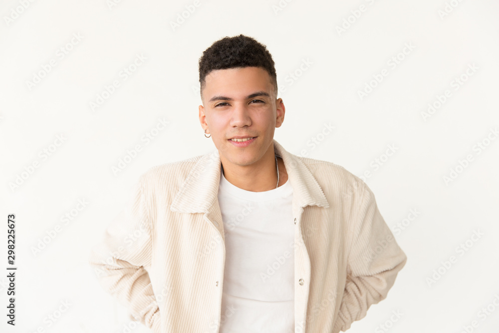 Handsome young man smiling at camera. Portrait of young man with curly hair and piercing standing with hands in pockets and looking at camera isolated on grey. Beauty concept Handsome young man smiling at camera. Portrait of young man with curly hair and piercing standing with hands in pockets and looking at camera isolated on grey. Beauty concept