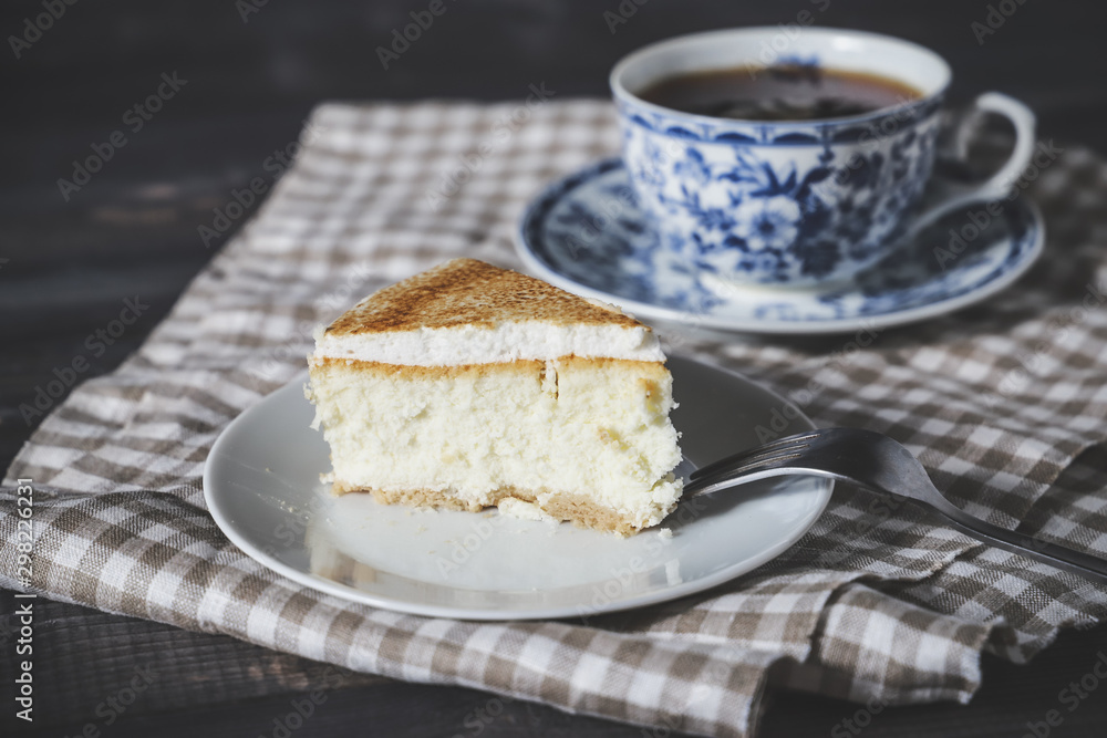 On a dark countertop, on a light checkered linen napkin, a white and blue patterned tea pair with tea and saucer with a piece of classic cheesecake and a fork. On a dark countertop, on a light checkered linen napkin, a white and blue patterned tea pair with tea and saucer with a piece of classic cheesecake and a fork.