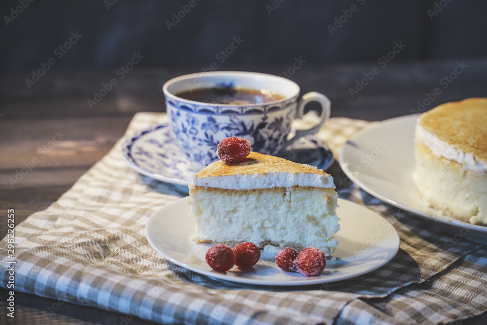 On a dark tabletop, on a light checkered linen napkin on a white saucer, a piece of cheesecake cake and a tea pair with tea On a dark tabletop, on a light checkered linen napkin on a white saucer, a piece of cheesecake cake and a tea pair with tea