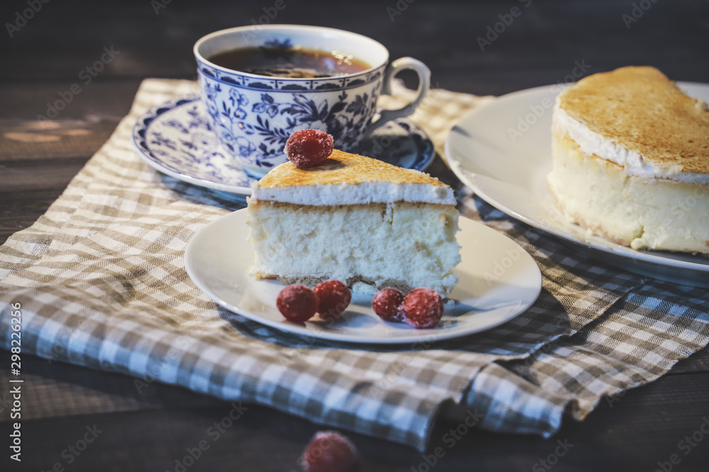 On a dark tabletop, on a light checkered linen napkin on a white saucer, a piece of cheesecake cake and a tea pair with tea