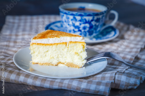 On a dark countertop  on a light checkered linen napkin  a white and blue patterned tea pair with tea and saucer with a piece of classic cheesecake and a fork.