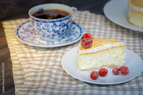 On the saucer is a piece of cheesecake cake and red berries, the whole cake on a white plate, next to it is a white tea pair with a blue pattern, everything is on a linen napkin.