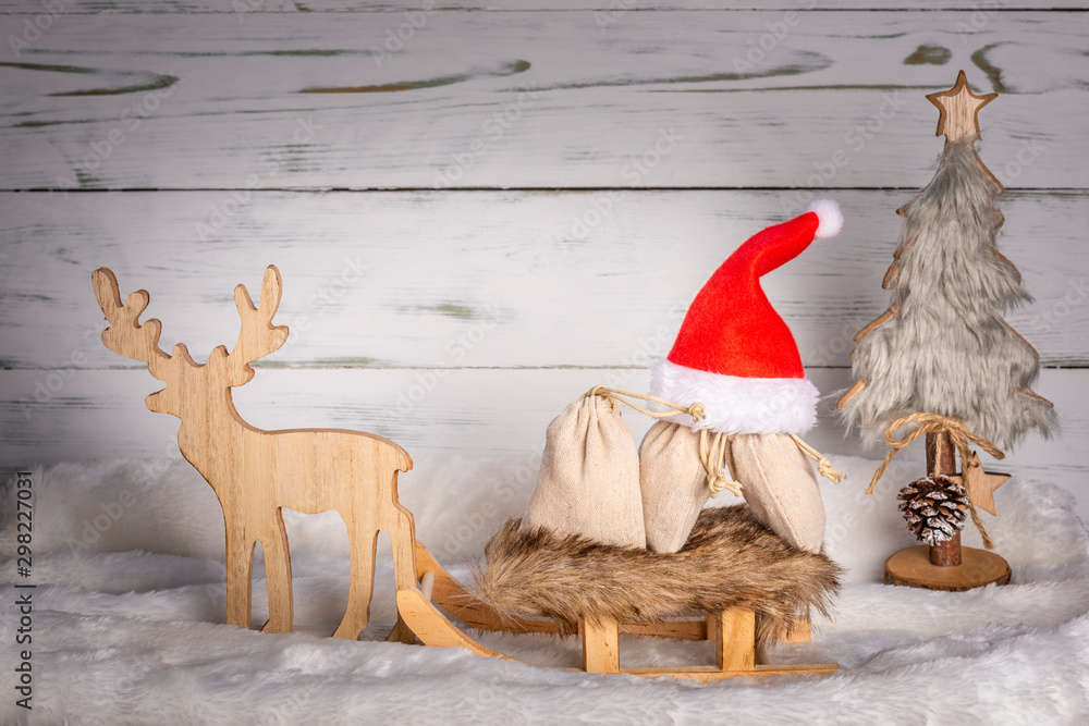 Obraz premium Weihnachtsmütze mit Schlitten, Rentier und Geschenke mit Weihnachtsbaum im Schnee vor einem Holz Hintergrund