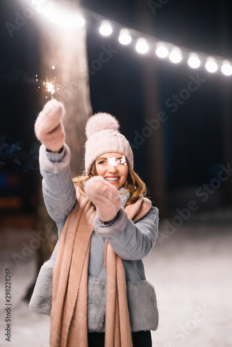 Beautiful girl with sparklers in hands. Happy winter time in the forest. The girl is dressed in a blue coat, in a light knitted hat, scarf and mittens. Festive garland lights. Christmas, new year.