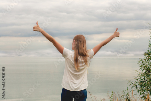 Beautiful girl on the lake. The view from the back. Hands up. Thumb up. Cloudy sky, sunset