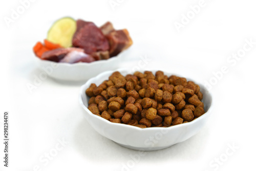 Concept for different dog feeding methods with bowl with dry food kibbles in front and blurry bowl with chunk of raw meat and vegetables in back isolated on white background