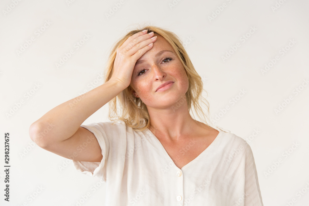 Frustrated woman with hand on forehead. Portrait of beautiful young blonde woman standing with hand on face and looking at camera isolated on grey background. Facial expression concept Frustrated woman with hand on forehead. Portrait of beautiful young blonde woman standing with hand on face and looking at camera isolated on grey background. Facial expression concept