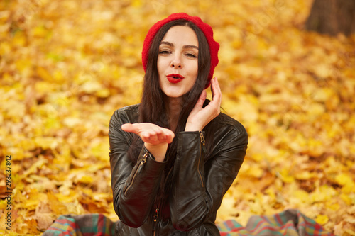 Close up portrait of elegant pretty brunette woman posing at autumn bright day in city park, wearing elegant red beret and stylish black leather jacket, sending air kiss to camera,warm autumn weather.