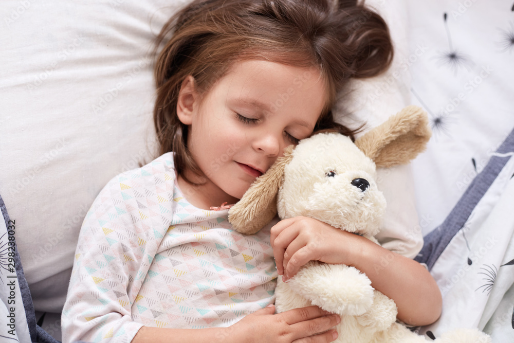 Close up portrait of young beautiful darkhaired girl, little princess with long hair, keeps eyes closed, child lies in bed, sleeping in linen with dondelion, hugging her favourite white soft dog toy.