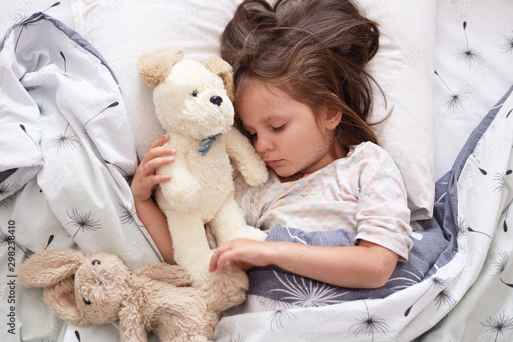 Close up portrait of beautiful cute little girl sleeping peacefully and hugging her stuffed toys in bed, charming kid with closed eyes, dark haired child relaxing at home in morning. Childhood concept