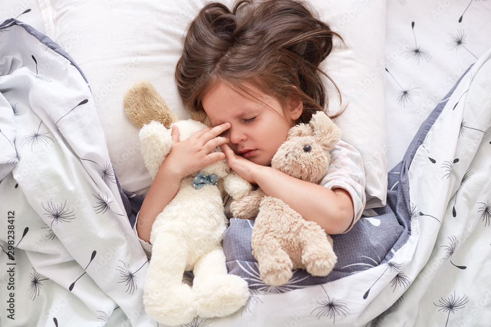 Close up portrait of cute little caucasian baby girl hugs soft teddy bear and dog toy. Portrait of sweetly sleeping toddler with pacifier, lying in bed with closed eyes. Concept of love and child care