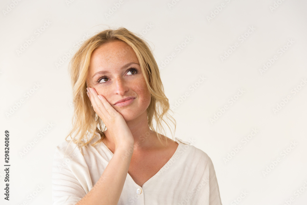 Pensive woman with hand on cheek. Beautiful thoughtful young woman standing with hand on face and looking up on grey background. Facial expression concept Pensive woman with hand on cheek. Beautiful thoughtful young woman standing with hand on face and looking up on grey background. Facial expression concept
