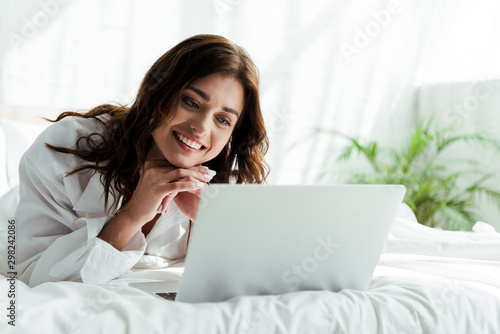 attractive woman in white shirt using laptop and smiling at morning