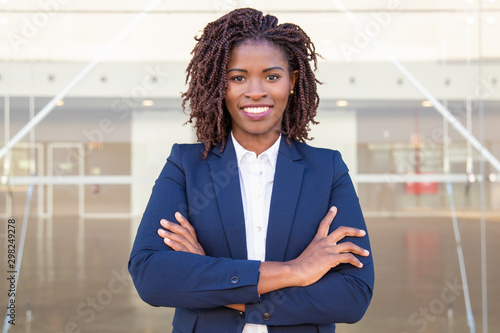 Happy successful business leader posing near outside. Young business woman with arms folded standing near glass wall, looking at camera, smiling. African American businesswoman concept