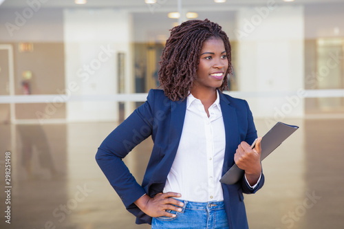 Cheerful successful manager with documents posing outside. Young black business woman standing at glass wall, holding folder, looking away, smiling. Successful professional concept photo