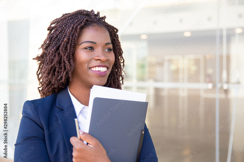 Happy cheerful office assistant carrying documents. Young African American business woman holding folder, standing outside, looking away, smiling, laughing. Assistant job concept
