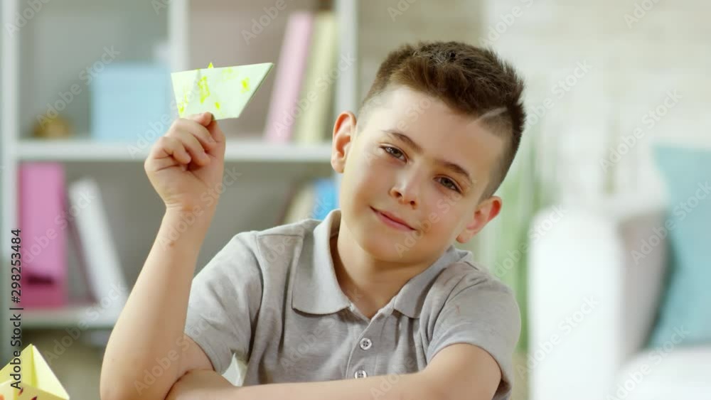 Joyous little caucasian boy holding handmade origami boat, looking at camera and smiling while posing at desk in art class Joyous little caucasian boy holding handmade origami boat, looking at camera and smiling while posing at desk in art class