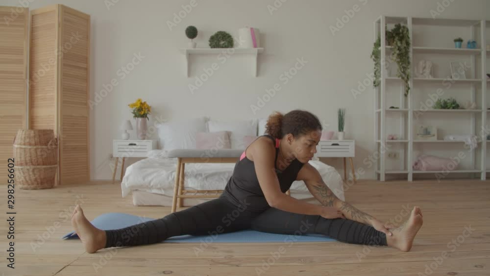 Motivated attractive young african american woman in sportswear doing stretching exercise while sitting on exercise mat in loft apartment. Sporty black woman practicing yoga warm up workout at home. Motivated attractive young african american woman in sportswear doing stretching exercise while sitting on exercise mat in loft apartment. Sporty black woman practicing yoga warm up workout at home.