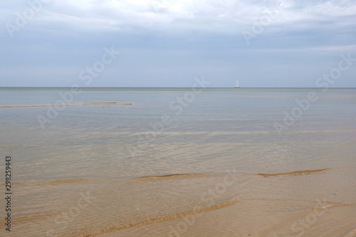 Fototapeta Naklejka Na Ścianę i Meble -  Sandy shoal of the cold Baltic Sea with cloudy weather on empty beach and alone yacht in drift on skyline