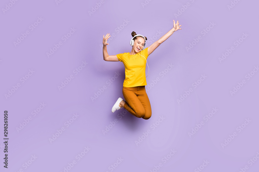 Full length photo of joyful lady jumping high listening modern playlist through cool earflaps dancing hip-hop wear yellow t-shirt trousers isolated pastel purple background