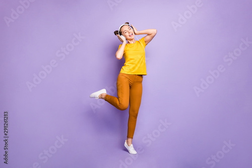 Fototapeta Naklejka Na Ścianę i Meble -  Full size photo of joyful teenager lady listening modern youngster playlist music through cool earflaps wear yellow t-shirt trousers isolated pastel purple background