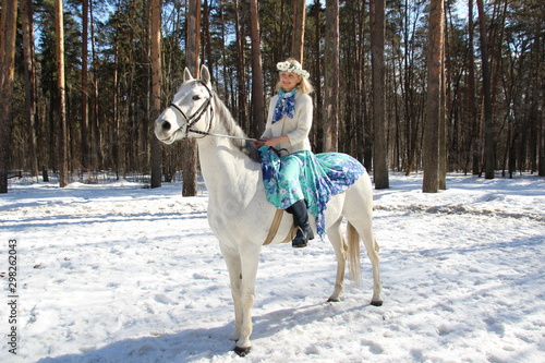 Beautiful happy young blond woman with flower crown in skirt on white horse in sunny winter day in the forest as a symbol of coming spring