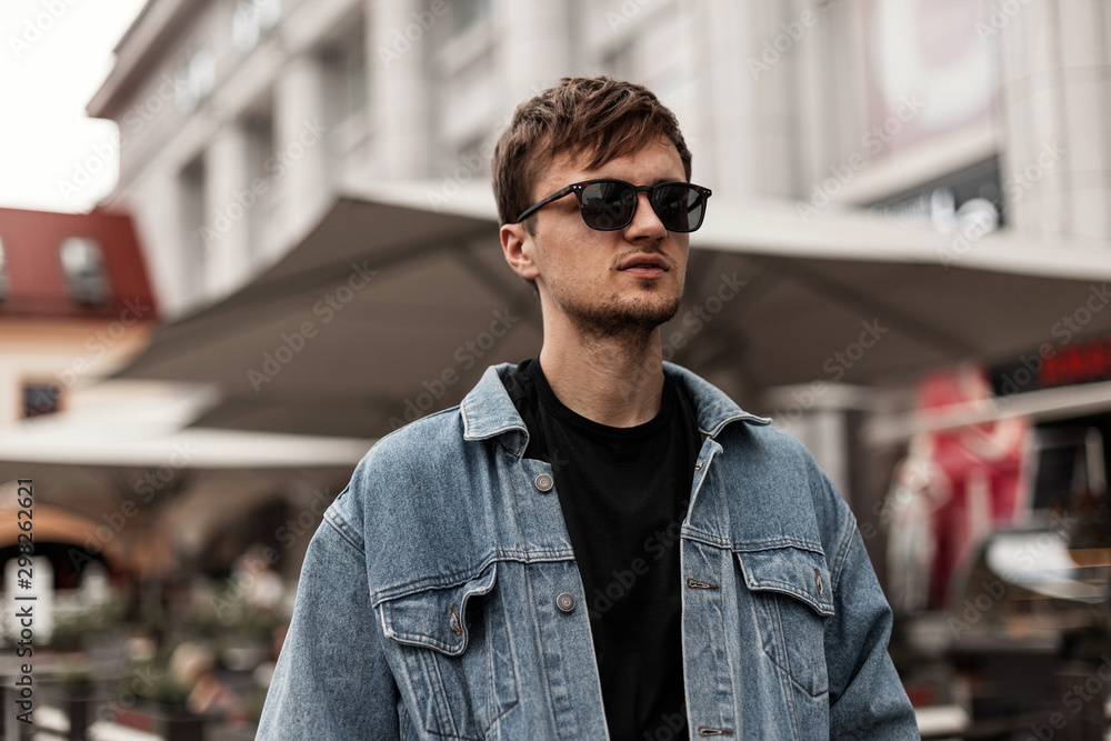 Cool nice young hipster man in a fashionable blue denim jacket in sunglasses stands on the street in the city. Handsome guy model with a stylish hairstyle rests outdoors. Fashion spring menswear. - obrazy, fototapety, plakaty 