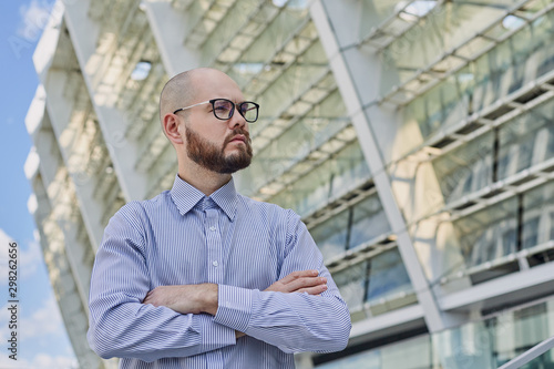 bald calm business man in a striped shirt with glasses stands with folded arms near surrender in the city