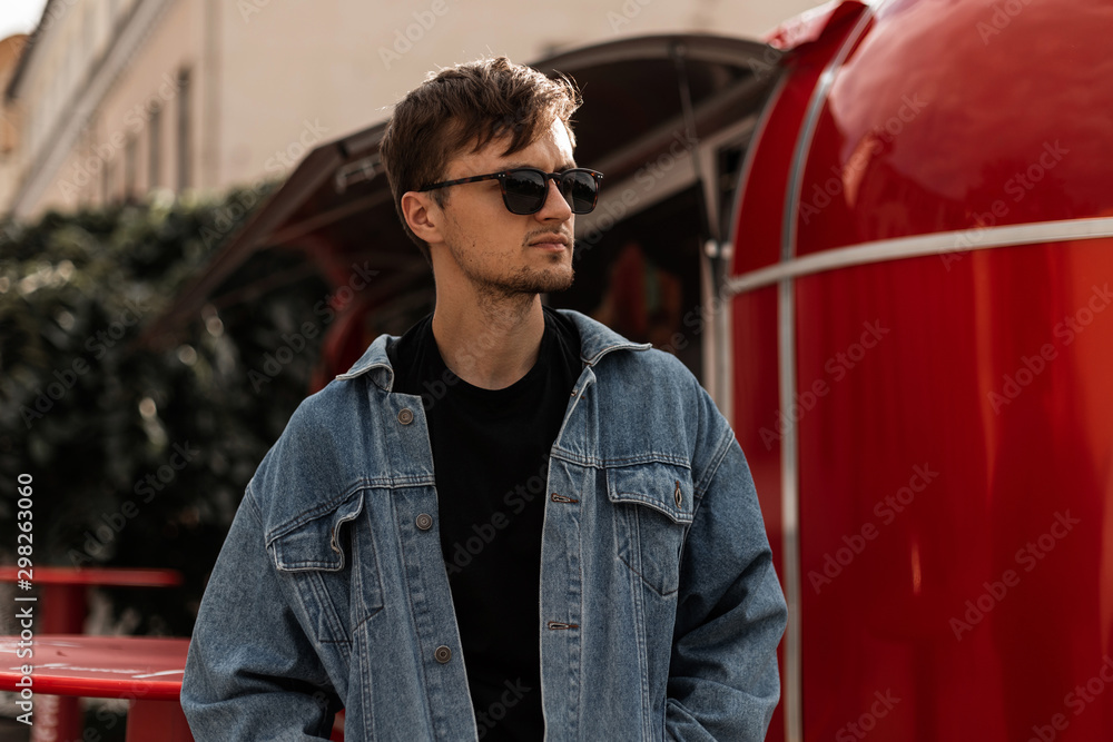 Stockfoto Nice stylish young hipster man in a denim jacket in a black  T-shirt in sunglasses with a hairstyle stands near a vintage metal red van.  Urban trendy guy model is resting