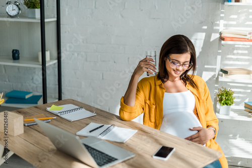 beautiful pregnant freelancer holding glass of water in office with home office