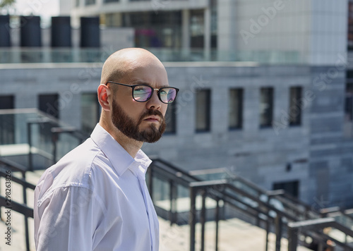 A bald young guy in glasses and with a dark beard on the street on the steps looking sideway against the background of a building in city. Worker, employee, employment. Business.