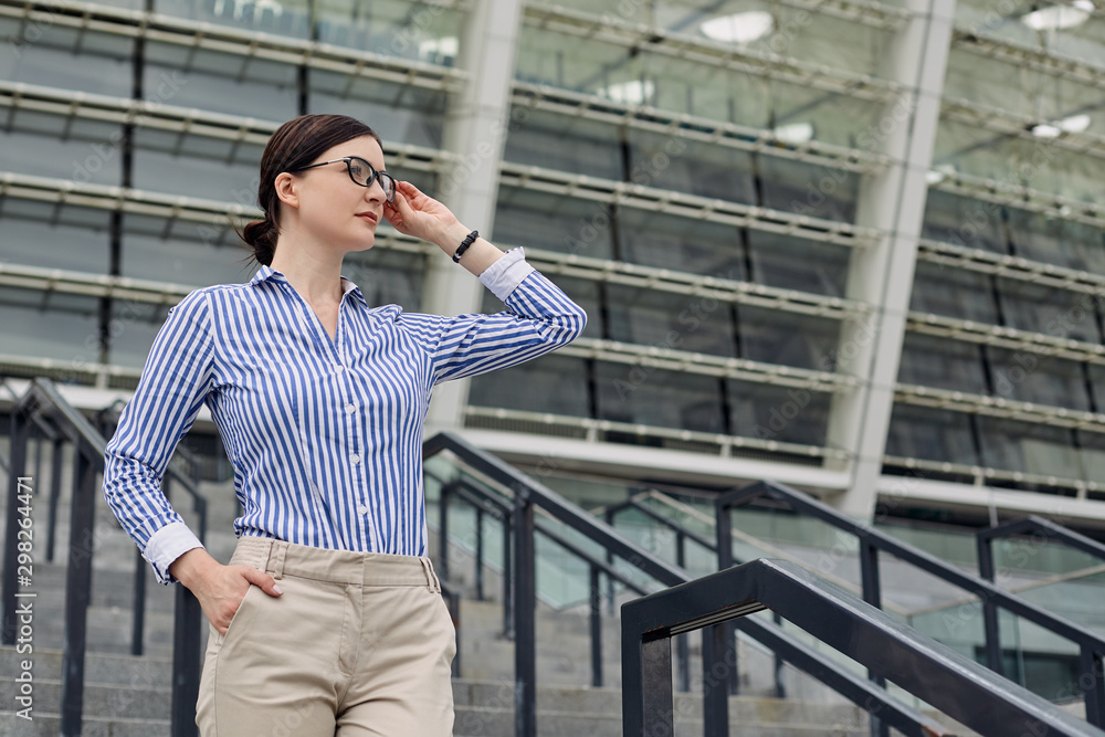 Business woman with brown hair holding on to glasses in a blue and white striped shirt with white cuffs on her sleeves against the background of city. Intelligent lady on steps. Business woman with brown hair holding on to glasses in a blue and white striped shirt with white cuffs on her sleeves against the background of city. Intelligent lady on steps.