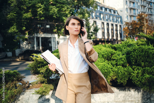 Young attractive businesswoman in beige suit with laptop thoughtfully talking on cellphone on street