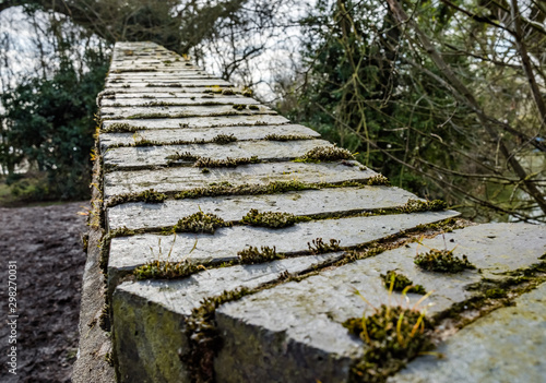 Close-up image of an old and weathered brick built foot bridge crossing. The surface of the walled area shows heaving weathering with moss growing between the brick joints as well signs of decay.