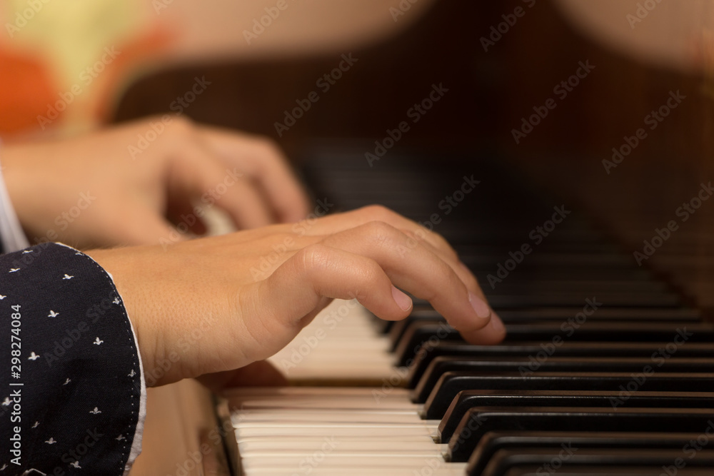 Close up side view of child hands playing the piano. Selective focus. Boy fingers and beautiful black shirt sleeves. Warm shade. Brown piano with black and white keys. Learning to play the piano