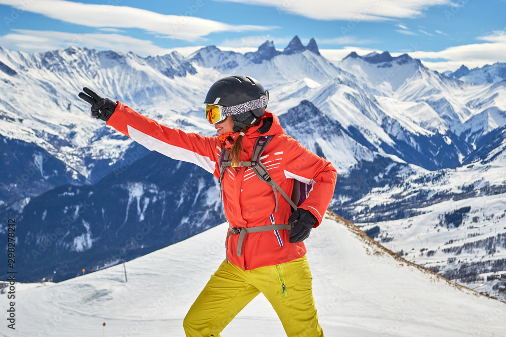 Woman skier smiling and posing at Les Sybelles ski domain, with Aiguilles d'Arves peaks in the background, on a sunny day with blue skies.